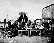 Men standing and sitting around two tables, facing the camera. A large tent behind them has a wooden sign that reads