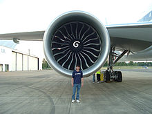 Aircraft engine, forward-facing view with a Boeing engineer in front to demonstrate the engine's size. The engine's large circular intake contains a center hub with a swirl mark, surrounded by multiple curved fan blades.