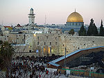 A large open area with hundreds of people, bounded by old stone walls. Beyond it are houses and a few trees, to the right is a mosque with large golden dome, and to the left &ndash; a minaret.
